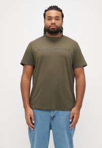 Oliivinvihreä puuvillainen t-paita, jossa on lyhyet hihat ja rintakehän keskellä tummansininen logopainatus. Yhdistetty vaaleansinisiin farkkuihin.