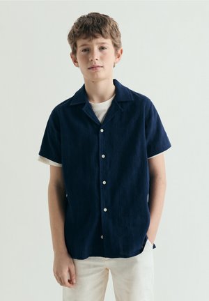 Chico con cabello corto y castaño que lleva una camisa de manga corta azul marino con botones sobre una camiseta blanca y pantalones de color claro, de pie contra un fondo liso.
