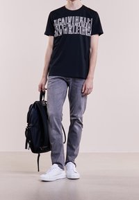 Persona que lleva una camiseta gráfica negra, jeans grises, zapatillas blancas, sosteniendo una mochila negra de un asa, de pie sobre un suelo claro.