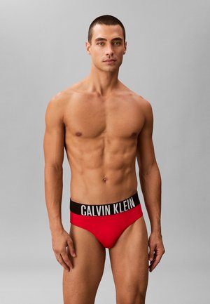 Männliches Model steht vor einfachem Hintergrund, trägt rote Calvin Klein Slips mit schwarzem Bund und weißem Logo, schaut nach vorne.