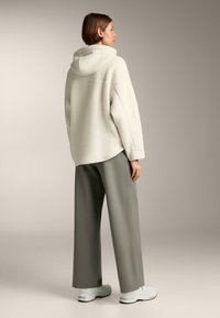 Giacca in pile color crema con cappuccio, taglio oversize e texture morbida, abbinata a pantaloni grigi larghi e sneakers chiare.