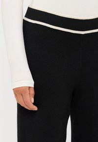 Černé kalhoty s jemnou texturou, s žebrovaným pasem a bílou linkou. Částečně viditelná ruka na nohavici. Dlouhý bílý rukáv podkal.