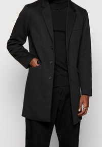 Cappotto nero con un design sartoriale, bottoni a un petto, rever a punta e vestibilità slim. Presenta una tasca laterale e dettagli minimi.