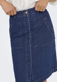 Jeans krilo v temno modri barvi s ravnim krojem, ki ima dva sprednja žepa in gumbasto zapiranje v pasu. Šivani detajli po celotni dolžini.