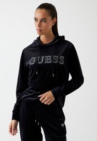 Vrouw met een staart die een zwarte velours hoodie en een broek draagt met een 'GUESS' gespeld logo op de borst en een bijpassend logo op de broek.