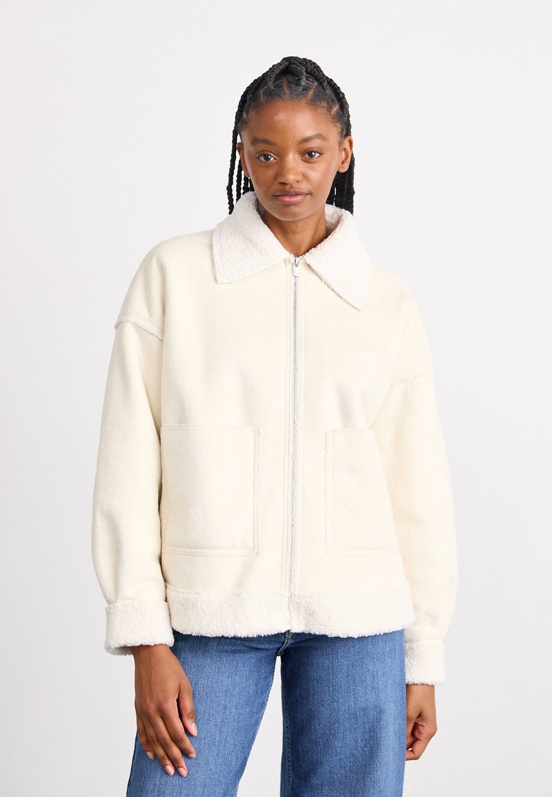 Veste en polaire couleur crème avec une finition douce et texturée, dotée d'un col, d'une fermeture éclair sur le devant et de deux grandes poches avant.