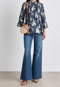 Blusa de estampado floral azul con detalles dorados, jeans acampanados y zapatos beige. Bolso beige con una correa marrón.