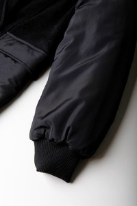 Svart bomberjacket med en glatt og blank nylonoverflate og ribbstrikkede mansjetter, med myk, teksturert stoff langs skuldrene.