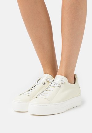 Białe niskie trampki z grubymi podeszwami noszone na gołych nogach na neutralnym tle.