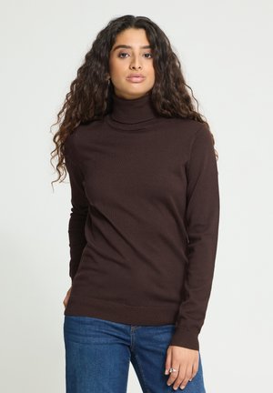 Jeune femme aux longs cheveux bouclés portant un pull à col roulé marron foncé et un jean bleu sur un fond clair.