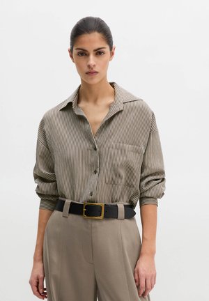 Kobieta o ciemnych włosach związanych w niski kok, ubrana w brązową i białą koszulę w pionowe paski, wpuszczoną w beżowe spodnie z czarnym paskiem z złotą klamrą.