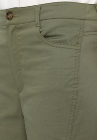 Pantalons en coton vert olive avec une texture lisse, dotés d'une poche frontale, de détails cousus et d'une fermeture à bouton marron.