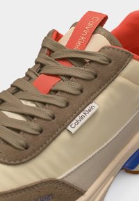 Μπεζ και καφέ αθλητικό παπούτσι Calvin Klein με ελιές κορδόνια, κόκκινη ετικέτα στη γλώσσα και μπλε λεπτομέρεια στη σόλα, κοντινή λήψη στο πλάι και την περιοχή των κορδονιών.