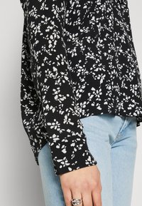 Svart bluse med hvitt blomstermønster, lange ermer og knappdetaljer ved mansjettene. Kombinert med lyseblå denimjeans. Myk stoffstruktur.