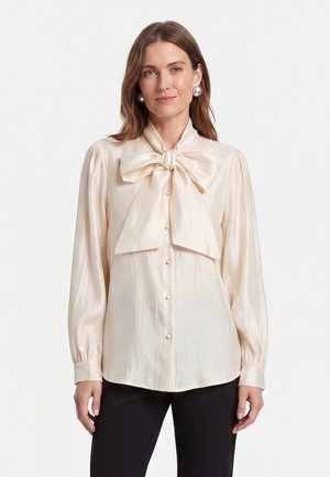 Vrouw draagt een crèmekleurige satijnen blouse met lange mouwen, parelknopen en een grote strik bij de hals, gecombineerd met een zwarte broek.