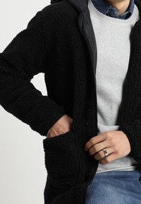 Veste en polaire noire avec une surface texturée, capuche et boutons. Les poches présentent des détails de couture visibles. Portée par-dessus un pull gris clair.