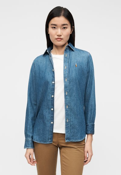 Sinine teksasärk, millel on nööbitav disain, pikad varrukad ja väike tikandiga logo rinnal. Kandmine valge särgi peal.