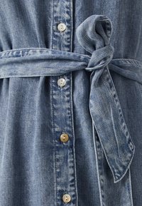 Vestido de ganga com um tom azul claro, apresentando um cinto amarrável, frente de botões e botões prateados. A textura é suave com um aspeto ligeiramente esbatido.