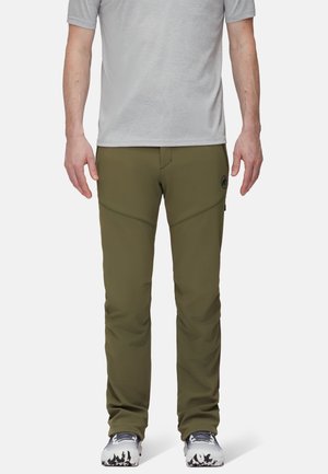 Man draagt olijfgroene softshellbroek, grijs T-shirt met korte mouwen en witte sportschoenen, staand tegen een effen achtergrond.