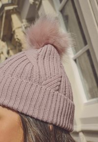 Kootud müts heleses roosas, millel on tekstuurne disain ribaga ja pehme vale karusnaha pom-pom otsas.