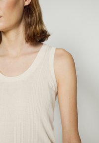 Femme aux cheveux bruns mi-longs portant un haut beige sans manches en maille côtelée, visible de l'épaule jusqu'au milieu du torse sur un fond neutre.