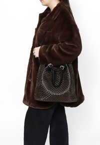 Bruine geweven emmer tas met leren handvatten, met een gestructureerd oppervlak en zilveren hardware-accenten. Draagt een bruin bontachtig jasje.