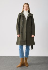 Zöld gyapjú peacoat dupla gombolással, fekete gombokkal és széles gallérral. Fehér garbó alatt viselve, kék farmerrel és barna csizmával párosítva.