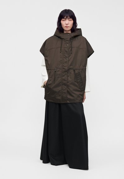 Femme portant une veste sans manches à capuche vert foncé par-dessus un haut blanc à manches longues et un pantalon large noir, debout devant un fond uni.