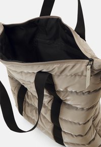 Sac fourre-tout matelassé beige avec une ouverture zippée, intérieur noir et poignées noires. Présente une petite étiquette avec le label "RAINS". Design texturé matelassé.