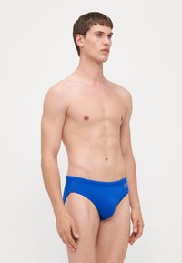 Blå svømmebokser med en glatt, blank tekstur, med en tapered cut og elastisk midje. Merkevaren logo er synlig på siden.