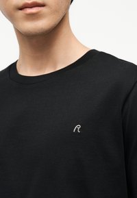 Κοντινό πλάνο ενός ατόμου που φοράει μαύρο μπλουζάκι με στρογγυλή λαιμόκοψη και ένα μικρό λευκό κεντητό γράμμα "R" στο στήθος.