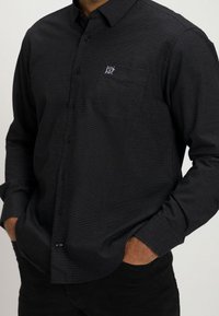 Homme portant une chemise noire ajustée à manches longues, texturée, avec un petit logo blanc brodé sur la poche poitrine.