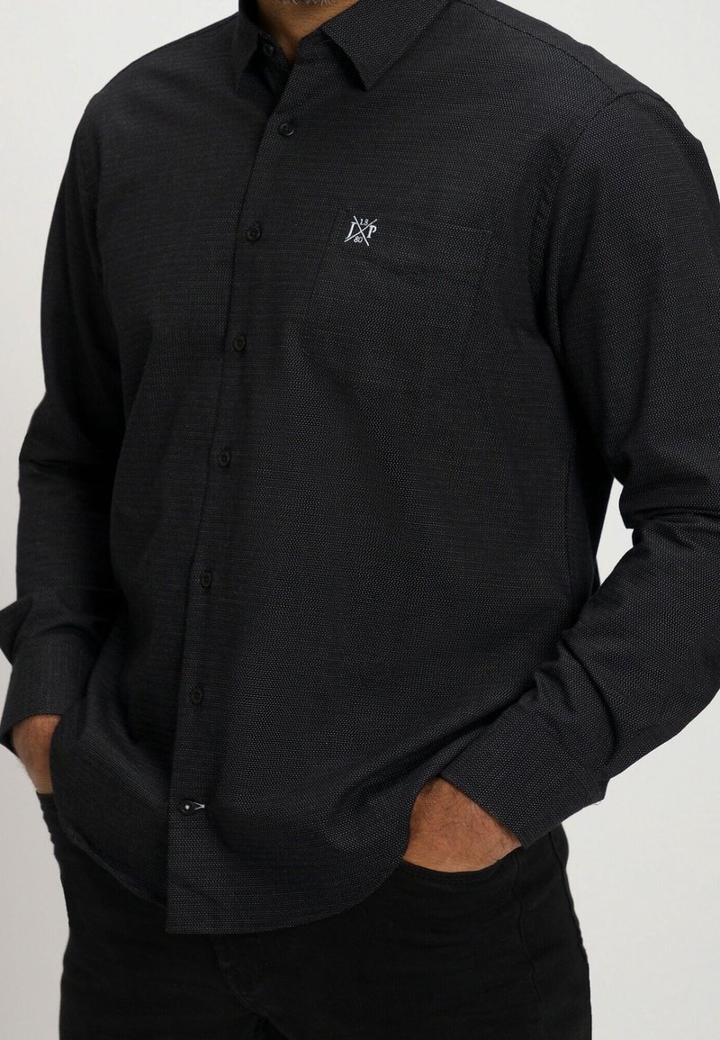 Homme portant une chemise noire ajustée à manches longues, texturée, avec un petit logo blanc brodé sur la poche poitrine.