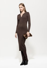 Braunes Strickkleid mit langen Ärmeln und Kragen, das einen subtilen Glanz aufweist. Kombiniert mit einer dunklen Fell-Clutch und kniehohen Lederstiefeln.