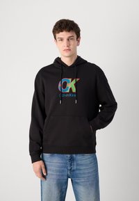 Fekete kapucnis pulóver pamutból, elülső kenguruzsebbel és húzózsinóros kapucnival. Piros és kék grafikával ellátott "CK" logóval rendelkezik.