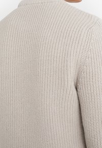 Zbliżenie jasnobeżowego swetra wykonanego na drutach, ukazujące teksturowany prążkowany wzór na plecach i obszarze ramion.