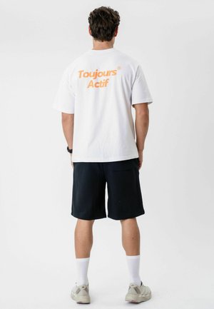 Mann steht mit dem Rücken zur Kamera, trägt ein weißes T-Shirt mit orangefarbenem "Toujours Actif"-Schriftzug, schwarze Shorts, weiße Socken und beige Sneakers vor weißem Hintergrund.