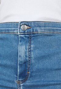 Siniset farkut, joissa on suora malli, metallinen nappikiinnitys ja näkyvät ompeleet saumoissa. Lähikuva vyötäröalueesta.