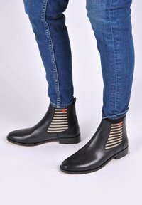Bottes noires en cuir à cheville avec des panneaux élastiques rayés et un détail en cœur rouge, associées à un jean bleu slim, sur fond blanc.
