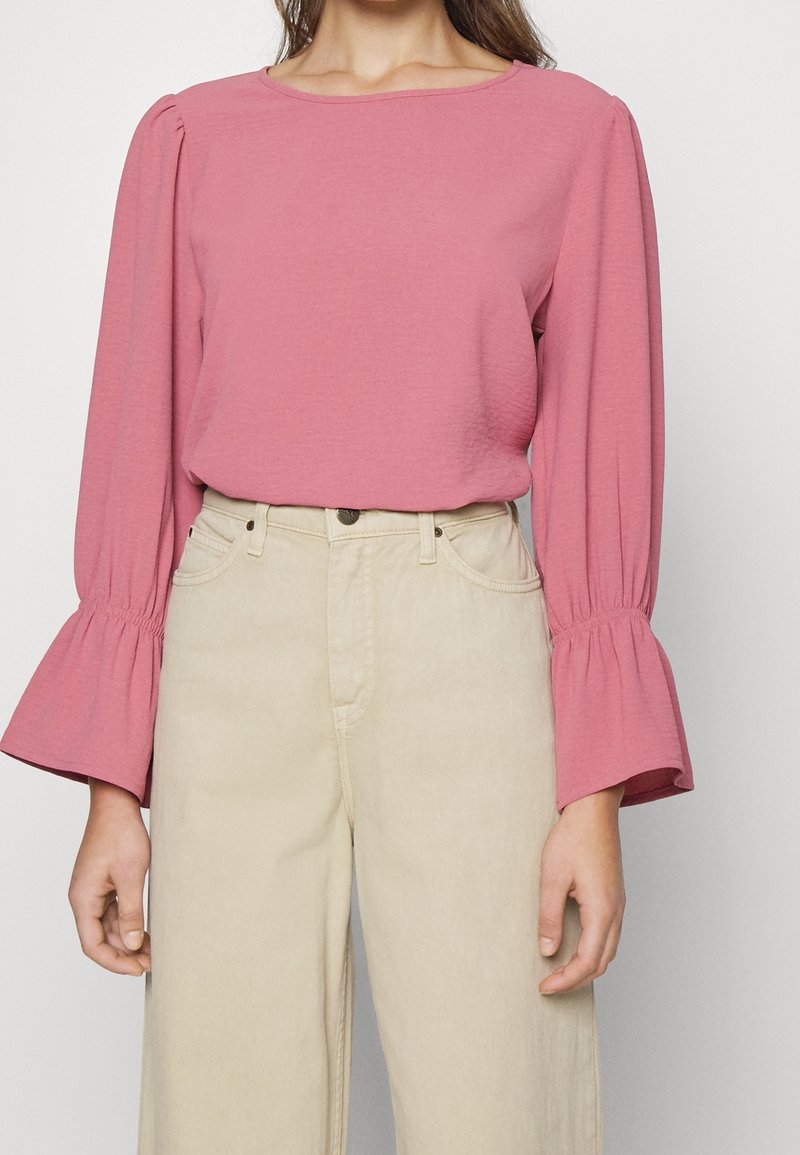 Femme portant un chemisier rose à manches longues avec des poignets évasés et un pantalon beige taille haute sur un fond uni.