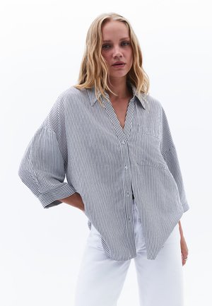 Camicia a righe bianca e nera con vestibilità rilassata, maniche ampie e tasca sul petto. Indossata sopra pantaloni bianchi.