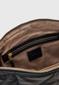 Fekete bőrtáska belseje, arany cipzárral és világosbarna béléssel. Tartalmaz egy cipzáras zsebet és egy fekete címkét domború felirattal.