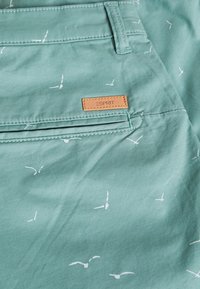 Tela azul verdosa con patrones de pájaros blancos y una etiqueta de cuero marrón de Esprit. Cuenta con un bolsillo con detalles de costura y una textura suave.