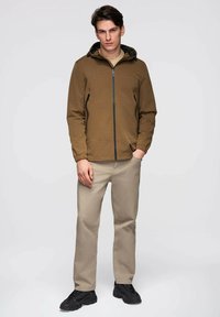 Jeune homme portant une veste à capuche marron, un pantalon beige et des baskets noires, se tenant avec une main dans la poche sur un fond uni.