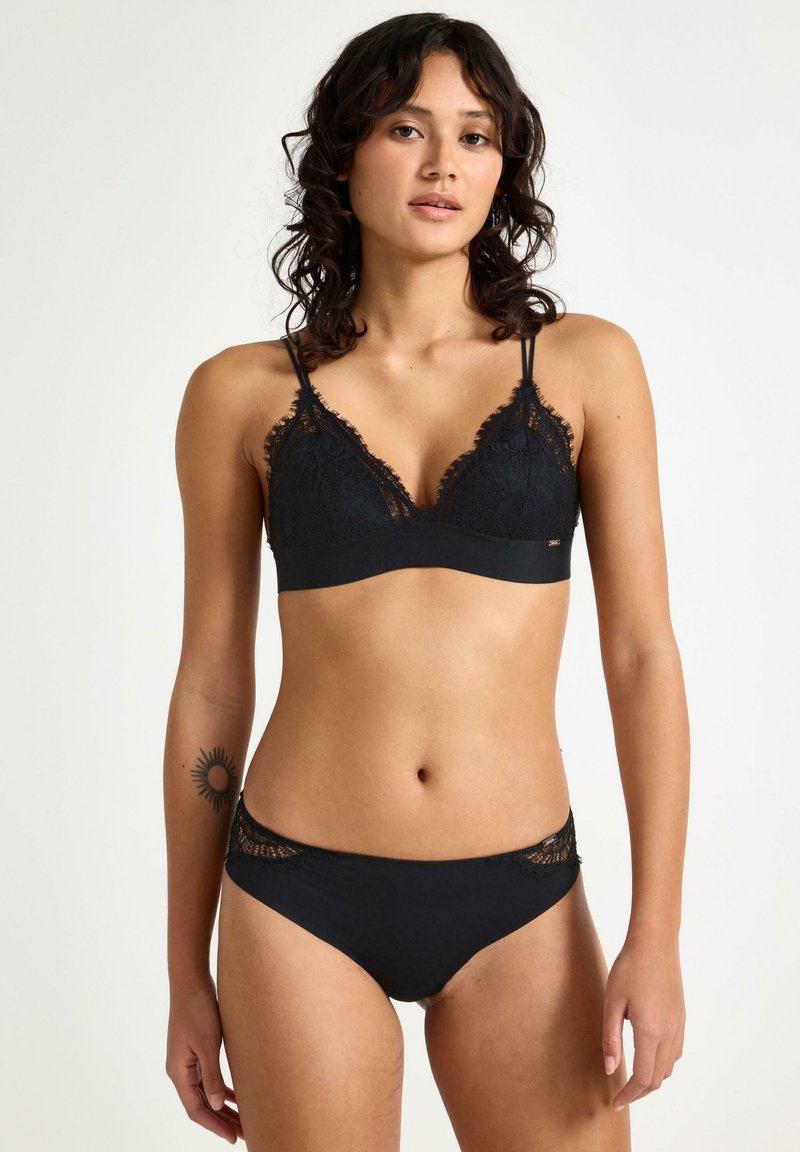 Musta pitsinen bralette säädettävillä olkaimilla, jossa on aaltoilevat reunat ja sileä vyötärönauha. Yhteensopivat mustat vähäpeitteiset panties, joissa on pitsireunus.
