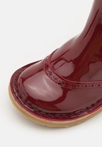 Bota impermeable roja brillante con punta redondeada, costuras intrincadas y una suela texturizada. Superficie lisa y reflectante con detalles sutiles.