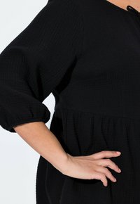 Black crinkled fabric top with a relaxed fit, featuring three-quarter puff sleeves and a gathered waistline. Accented with a small tie.