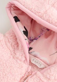 Primo piano di una giacca in pile teddy rosa con fodera interna stampata floreale e cursore della cerniera in oro rosa.