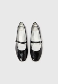 Bailarinas de charol negro con punteras cuadradas, un adorno de hebilla plateada y un acabado brillante. Interior suave y forrado visible.