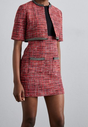 Mujer que lleva un vestido de tweed texturizado rojo y negro con una chaqueta corta a juego, con ribete negro y dos bolsillos delanteros en la falda.